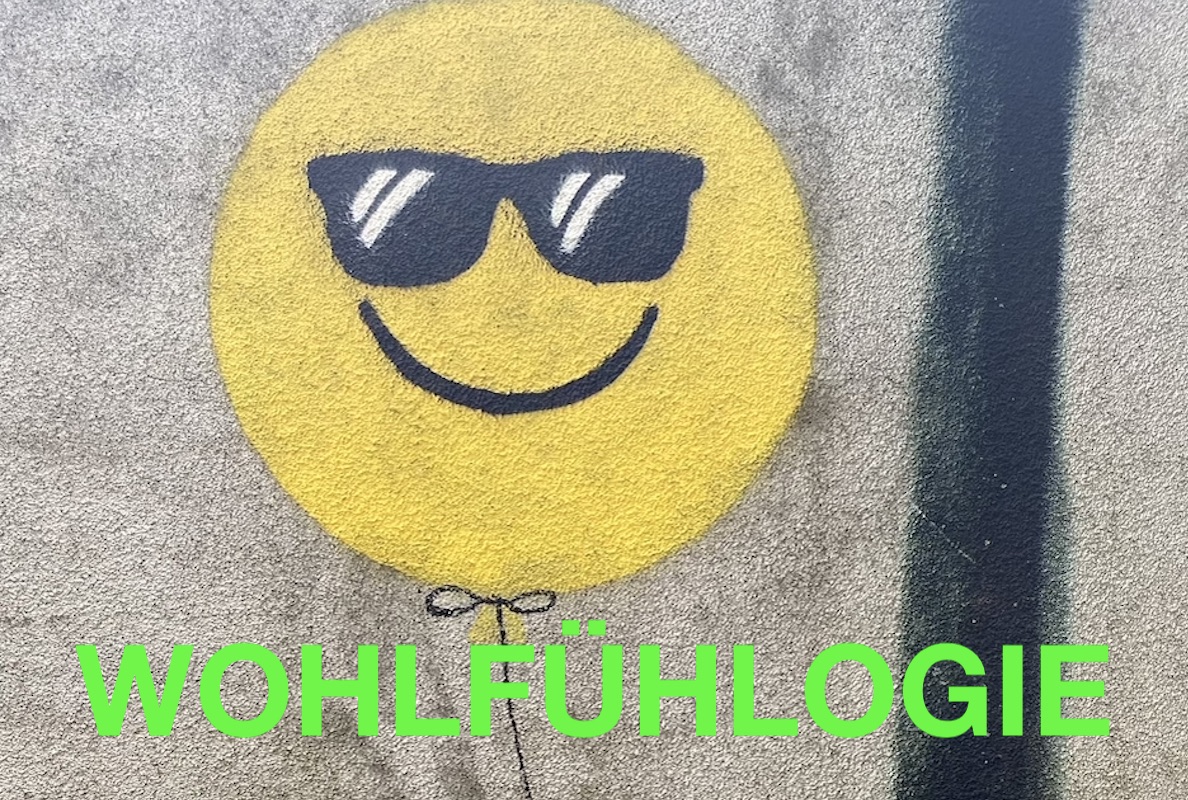 Grafitti eines gelben Luftballons mit schwarzer Sonnenbrille und Smiley-Lächeln, darunter das Wort Wohlfühlogie